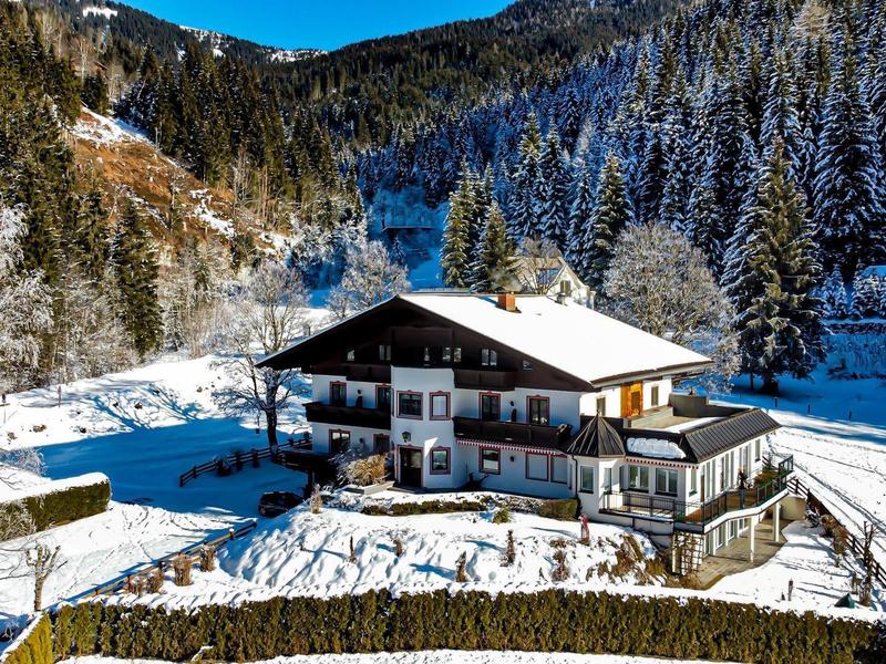 Starý chalet v zasněženém údolí obklopeném lesem a horami v pozadí.