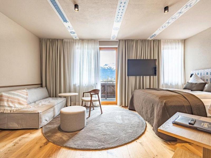 Heldere hotelkamer met bed, bank, tv en groot raam met uitzicht op bergen.