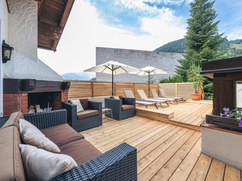 Gezellig houten terras met zithoeken, parasols en ligstoelen tegen een bergachtergrond.