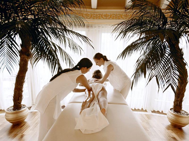 Two women in white dresses help a bride adjust her wedding dress, surrounded by palm trees.