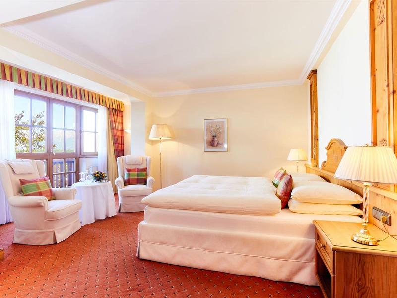 Bright hotel room with two beds, armchairs, carpet, and large window with outdoor view.