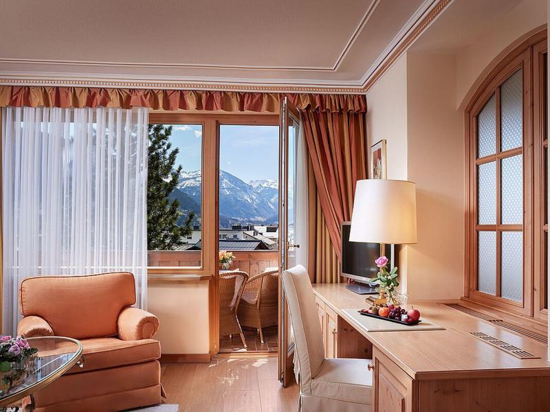 Brightly furnished hotel room with armchairs, desk, and mountain view through the window.