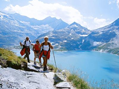 Tre escursionisti su un sentiero con un lago di montagna e cime innevate sullo sfondo.