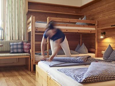 Gemütliches Zimmer mit Holzwänden, Etagenbett und Einzelbett mit karierten Decken und Kissen.