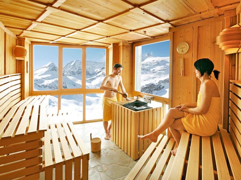 Innenansicht einer Holzsauna mit Panoramafenster und Blick auf verschneite Berge, zwei Personen sitzen drin.