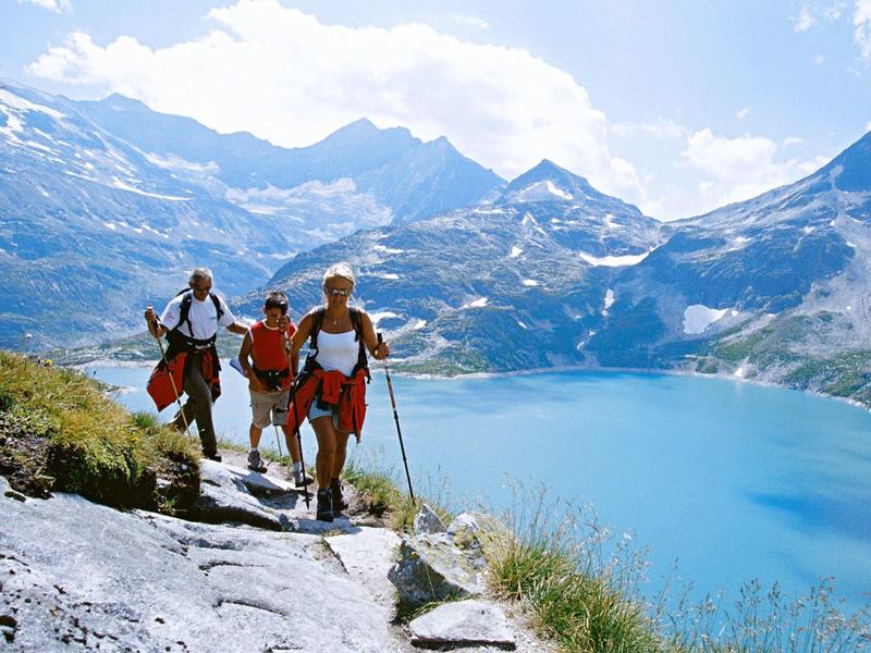 Vier Wanderer auf Bergweg mit blauen Bergsee und schneebedeckten Gipfeln im Hintergrund.