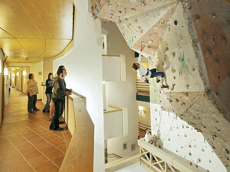 Innenbereich einer Kletterhalle mit Kletterwand und Menschen, die von einem Balkon zuschauen.