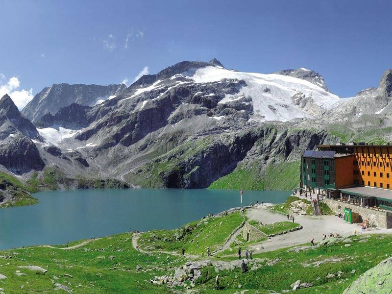 Bergsee mit grünem Ufer, schneebedeckten Gipfeln und einem gelben Berghaus an einem sonnigen Tag.