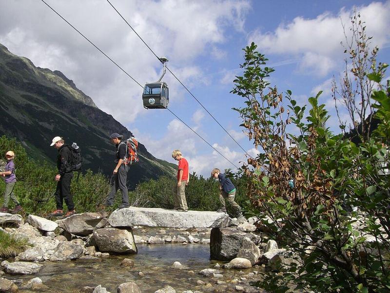Berglandschaft mit Seilbahn, Wanderern auf Felsen und bewölktem Himmel im Hintergrund.