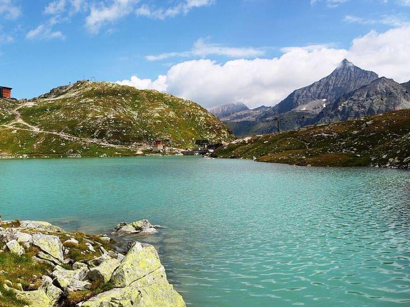 Bergsee mit türkisfarbenem Wasser, umgeben von Felsen, grünen Hügeln und einem Gebäude auf dem Gipfel.