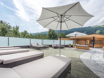 Moderní venkovní prostor s lehátky, slunečníkem a dřevěnou chatkou před zalesněnými kopci