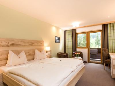 Helles Schlafzimmer mit Doppelbett, Holzdekoration, Sesseln, großem Fenster und Zugang zum Balkon.