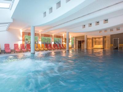 Interiér osvětleného vnitřního bazénu s lehátky a velkými okny.