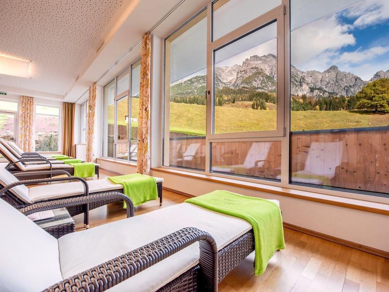 Heller Ruheraum mit weißen Liegen vor großen Fenstern und Bergblick, Holzboden und grünen Beistelltischen.