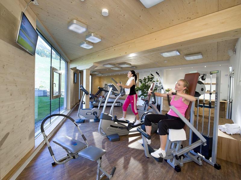 Hell gestalteter Fitnessraum mit Holzdecke, großen Fenstern und verschiedenen Trainingsgeräten.