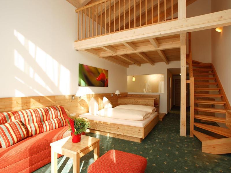 Gemütliches Zimmer mit Holzmöbeln, rotem Sofa, grünem Teppich und offener Holztreppe.