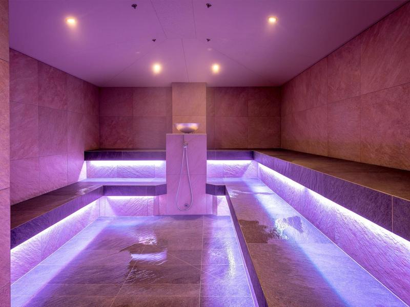 Moderner Wellnessraum mit lila Beleuchtung, Sitzbänken aus Holz und Mittelstele mit Wasserspender.
