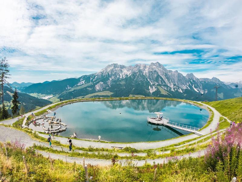 Rundlicher Bergsee mit Wegen, umgeben von grünen Hügeln und Bergen unter bewölktem Himmel.