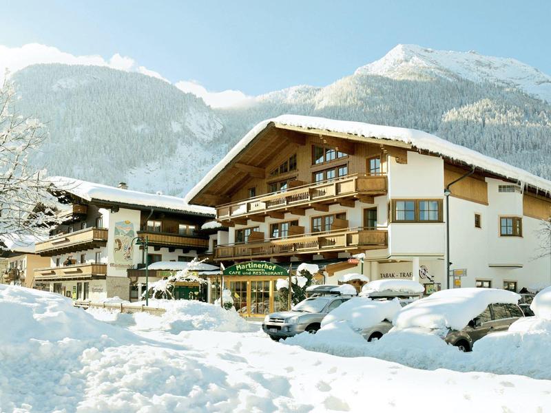 Bâtiment hôtelier hivernal devant des montagnes enneigées avec des voitures couvertes de neige au premier plan.