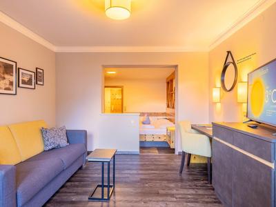 Habitación de hotel luminosa con sofá, escritorio, TV y vista al dormitorio con dos camas.