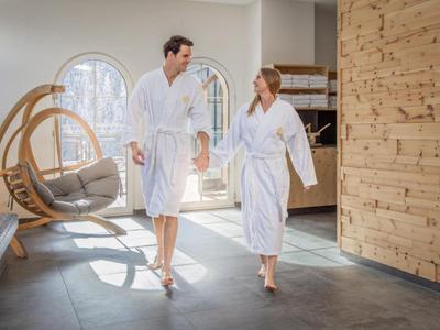 Una pareja con albornoces blancos camina tomada de la mano en un área de spa moderna y luminosa.