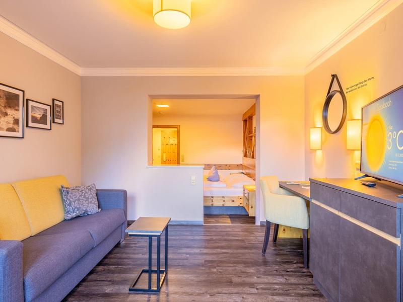 Habitación de hotel luminosa con sofá, escritorio, TV y vista al dormitorio con dos camas.