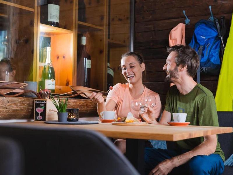 Dos personas ríen y beben en un acogedor restaurante de madera con chaquetas coloridas de fondo.