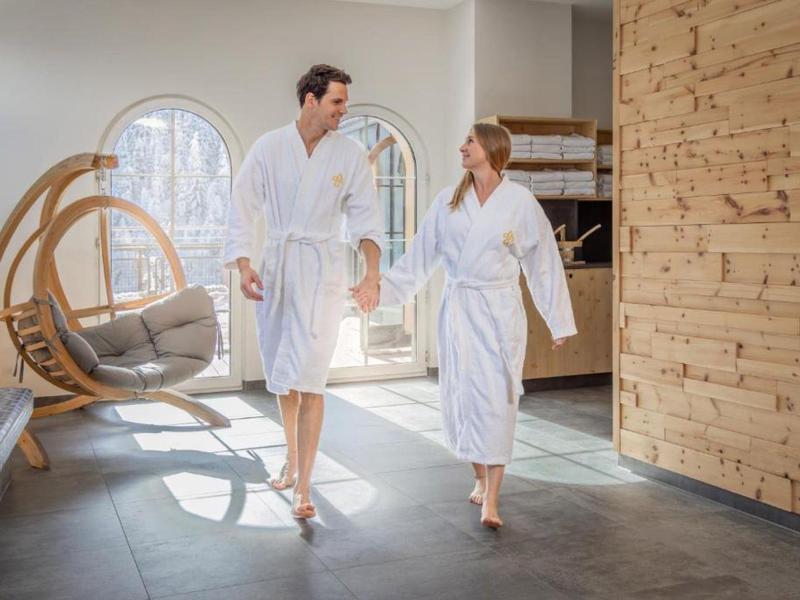 Una pareja con albornoces blancos camina tomada de la mano en un área de spa moderna y luminosa.
