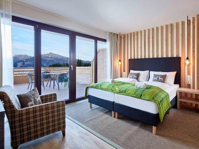 Modern hotel room with large bed, armchairs, and balcony overlooking mountains.