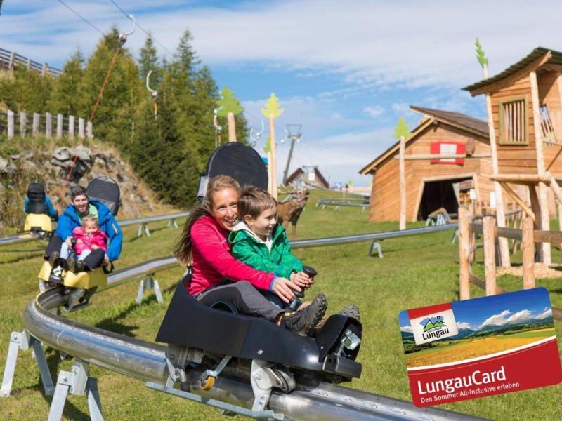 Two children are riding a summer toboggan run in a wooded mountain area.