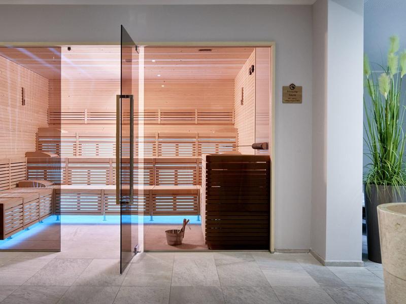 Modern sauna area with glass door, wooden benches, and bucket, surrounded by tiled floor.