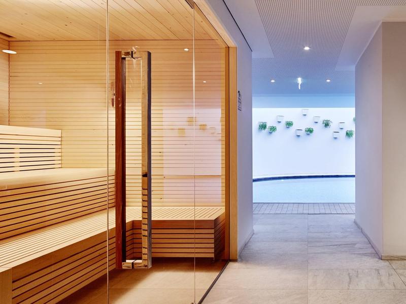 Modern sauna room with wooden benches and glass wall next to a brightly lit corridor.