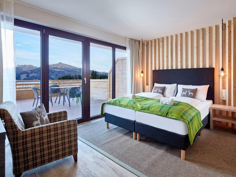 Modern hotel room with large bed, armchairs, and balcony overlooking mountains.