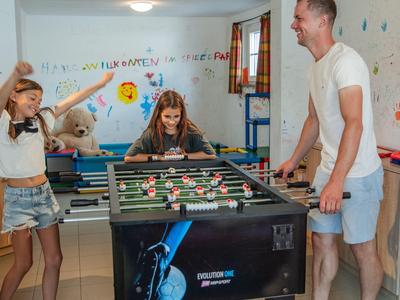 Dvě dívky a muž hrají stolní fotbal v jasné a barevné místnosti.