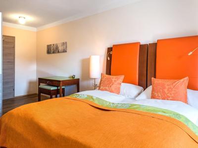 Světý hotelový pokoj s manželskou postelí, oranžovými přikrývkami a moderním dřevěným nábytkem.