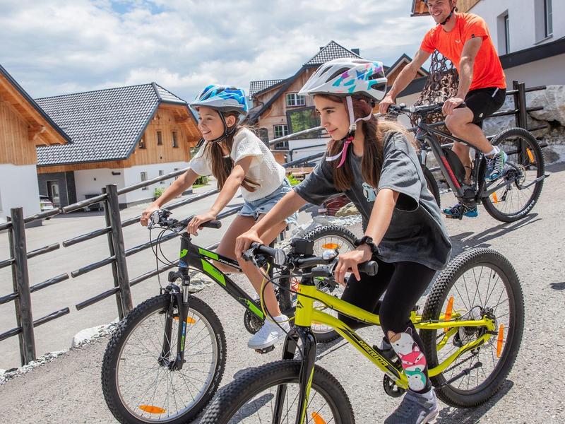 Tři lidé jedou na kolech v přilbách podél silnice s dřevěnými budovami v pozadí.
