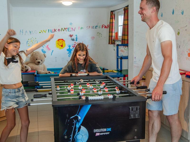 Dvě dívky a muž hrají stolní fotbal v jasné a barevné místnosti.
