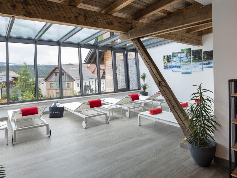 Světla hotelová hala s velkými okny, bílými židlemi a červenými polštáři s výhledem na hory.