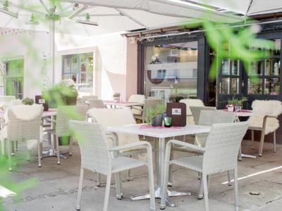 Area esterna vuota di un caffè con tavoli e sedie bianchi sotto ombrelloni.