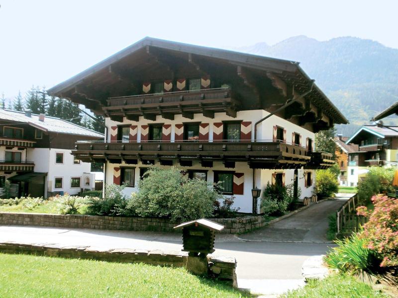 Maison alpine traditionnelle avec balcons en bois et fleurs, entourée de montagnes et d'espaces verts.