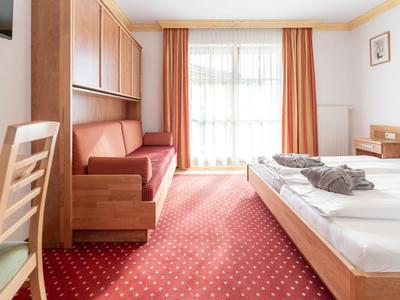 Světly hotelový pokoj s manželskou postelí, lavicí, psacím stolem a velkými okny se záclonami.