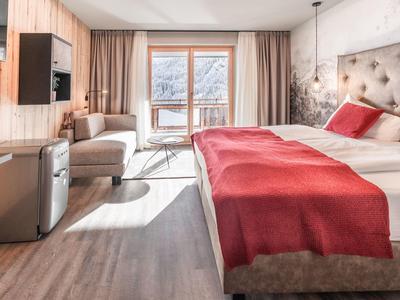 Světý hotelový pokoj s manželskou postelí, červeným povlečením, pohovkou a balkonem s výhledem na hory.