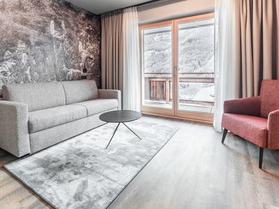 Světý obývací pokoj s pohovkou, červeným křeslem, kulatým stolem a velkými okny se závěsy.