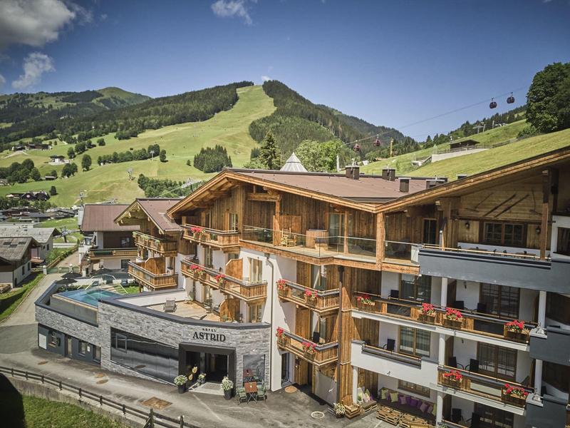 Horský hotel se dřevěnými balkony, vířivkou a zelenými kopci za jasné oblohy.