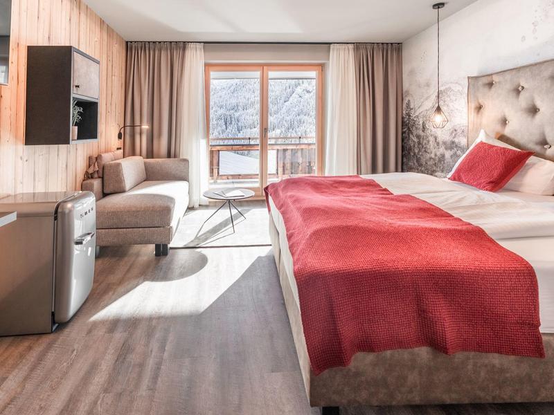 Světý hotelový pokoj s manželskou postelí, červeným povlečením, pohovkou a balkonem s výhledem na hory.