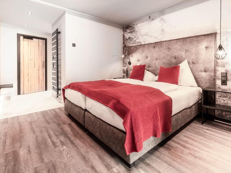 Moderní hotelový pokoj s manželskou postelí, červenými přikrývkami a dřevěnou podlahou.