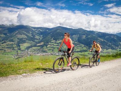 Zwei Radfahrer auf einem Schotterweg mit grünen Bergen und blauem Himmel im Hintergrund.