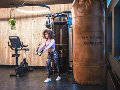 Frau trainiert mit Widerstandsband im Fitnessraum neben Boxsack und Heimtrainer.