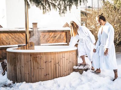 Paar in Bademänteln steigt in einen Holz-Whirlpool im Schnee im Freien.