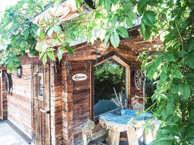 Kleines Holzhaus mit Terrasse und Whirlpool, umgeben von grünen Blättern.